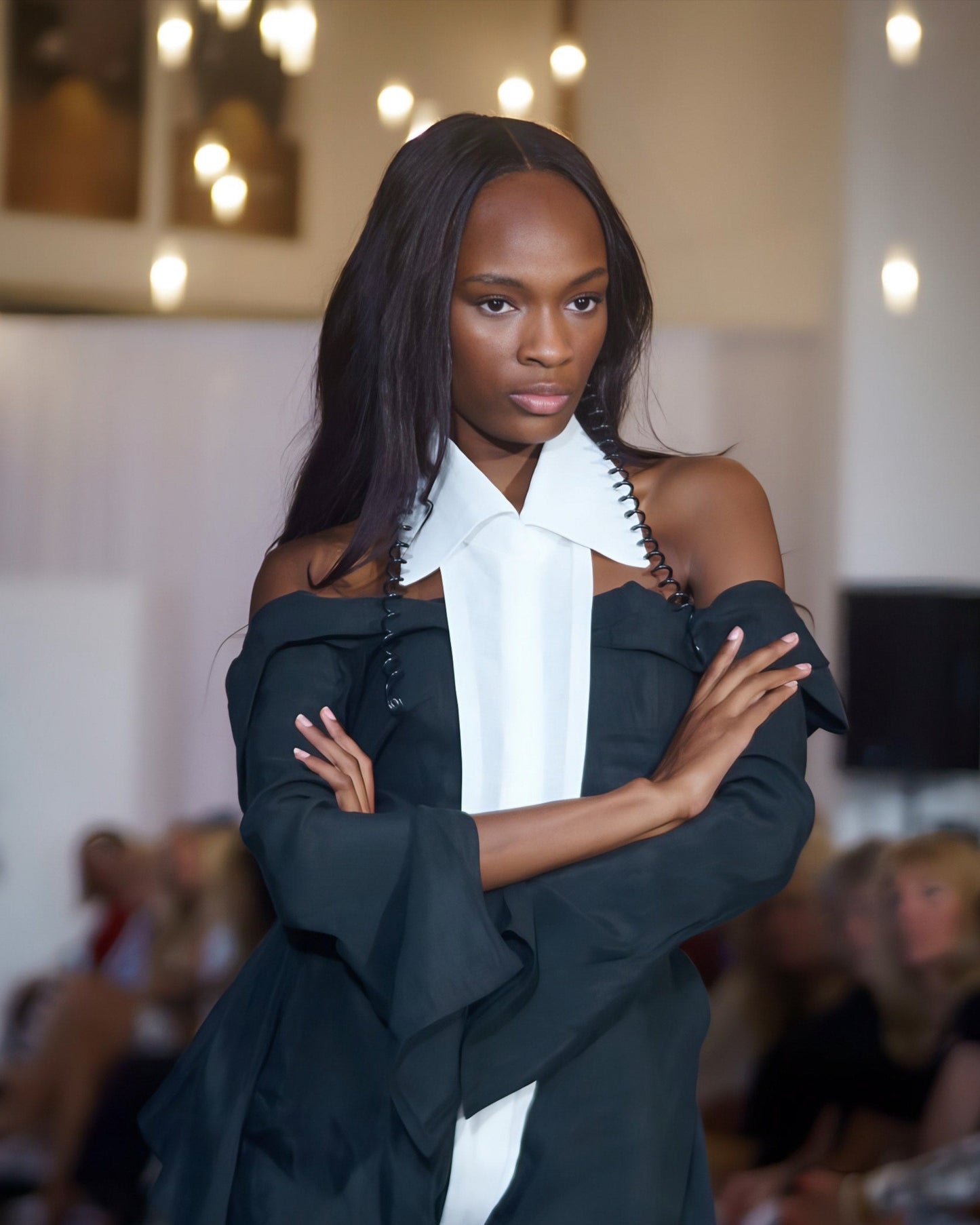 Model wearing a black outfit with a white collar in a fashion show setting