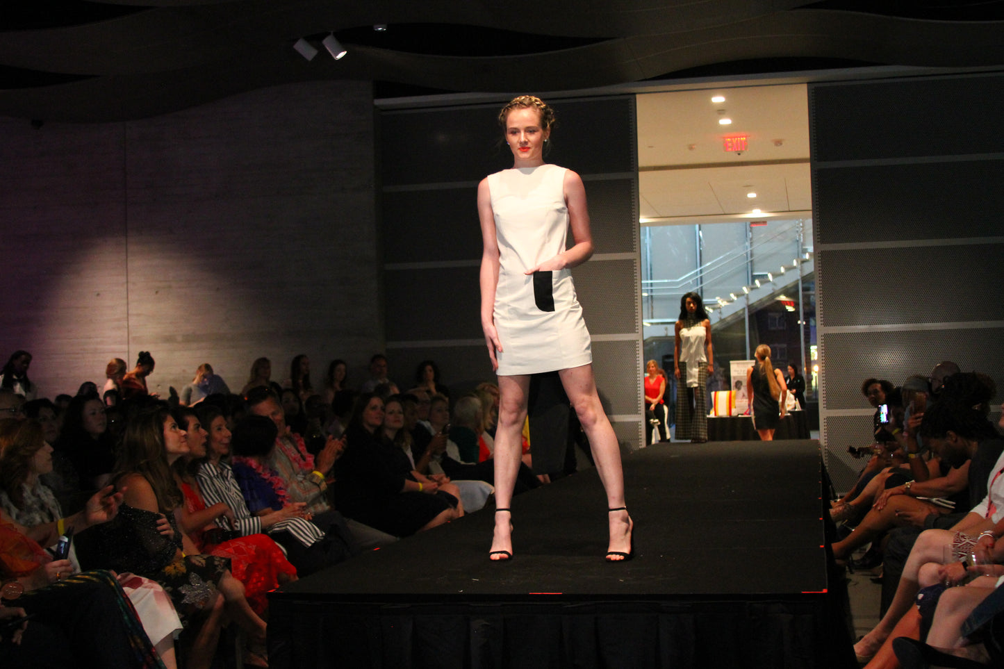 Anastasi Fashion design – Eclipse Dress (Front): A female model wearing a white twill dress with black zippered center back seams, an eggshell white body, and black front pocket. The dress is presented on a runway with an audience in the background.