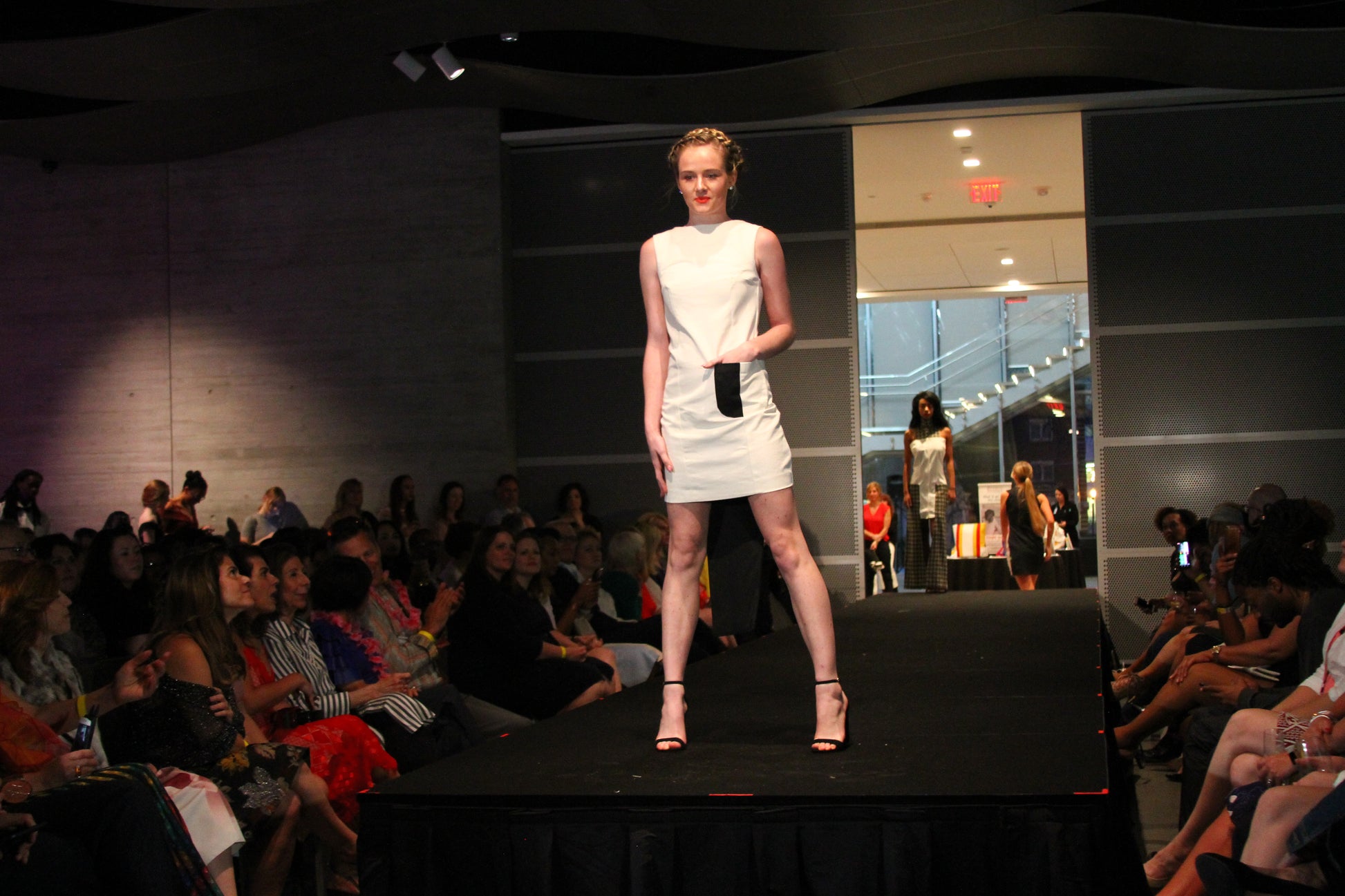 Anastasi Fashion design – Eclipse Dress (Front): A female model wearing a white twill dress with black zippered center back seams, an eggshell white body, and black front pocket. The dress is presented on a runway with an audience in the background.