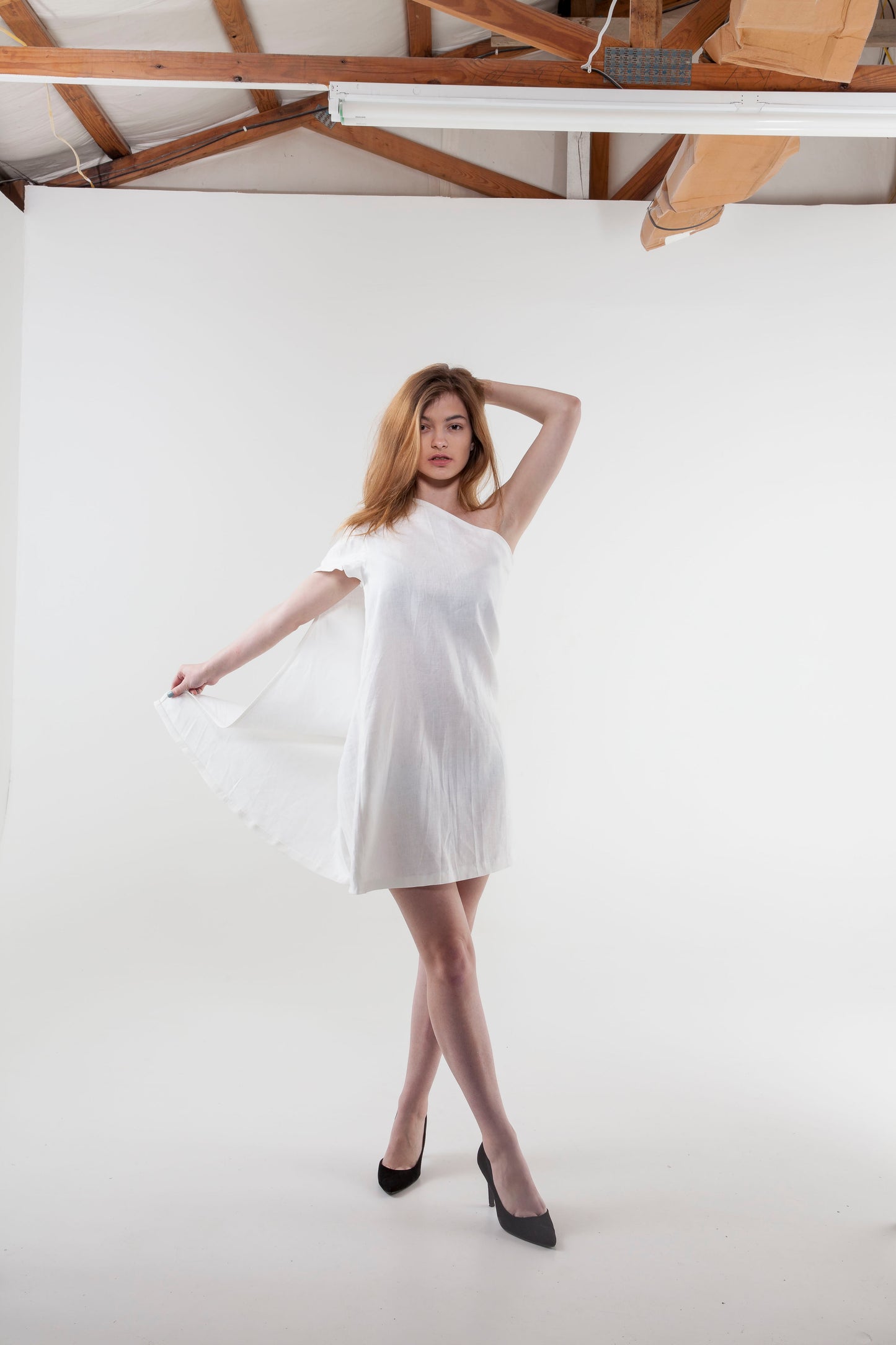 Anastasi Fashion design – Off the Shoulder Cape Dress (Front Stance): A woman modeling an off-the-shoulder white linen cape dress with a flared skirt, posing against a white background.